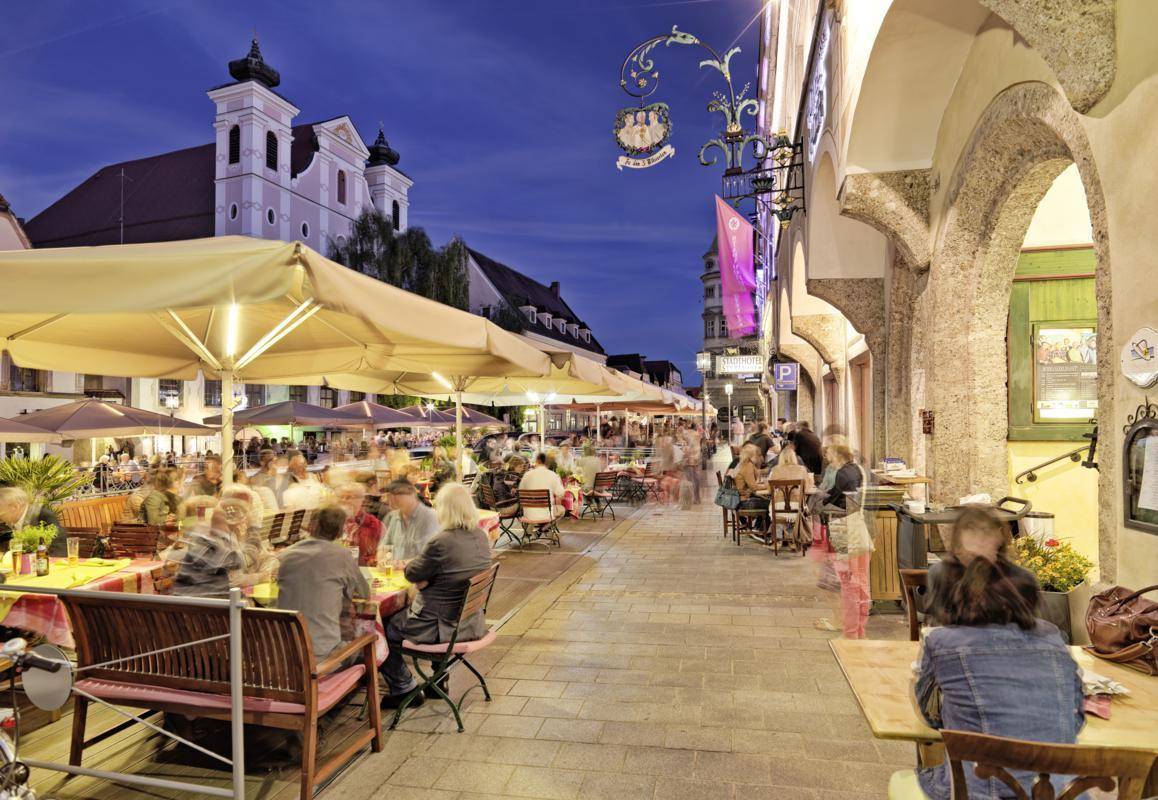 Belebte Außenterrasse eines Restaurants am Abend mit vielen Gästen und historischer Architektur im Hintergrund.