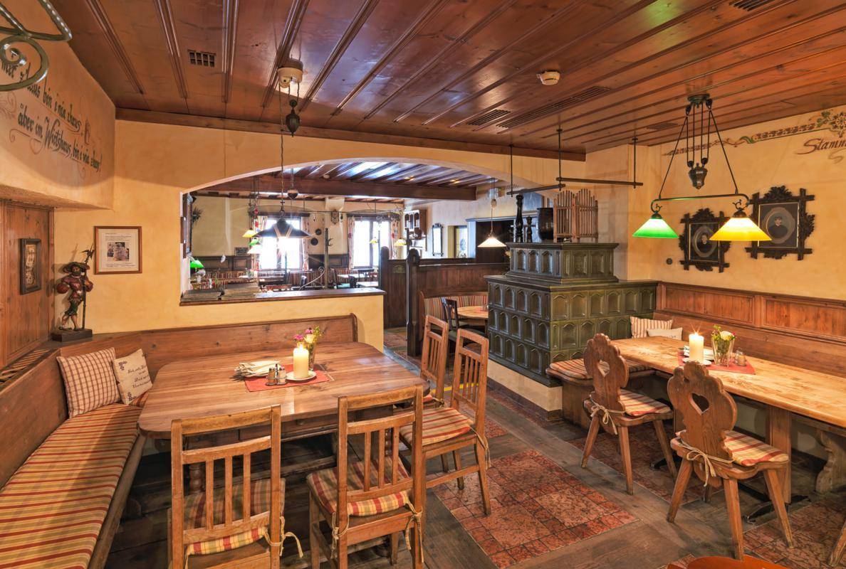 Rustikaler Restaurantbereich mit Holztischen, Bänken und einem traditionellen Kachelofen, gemütlich eingerichtet.