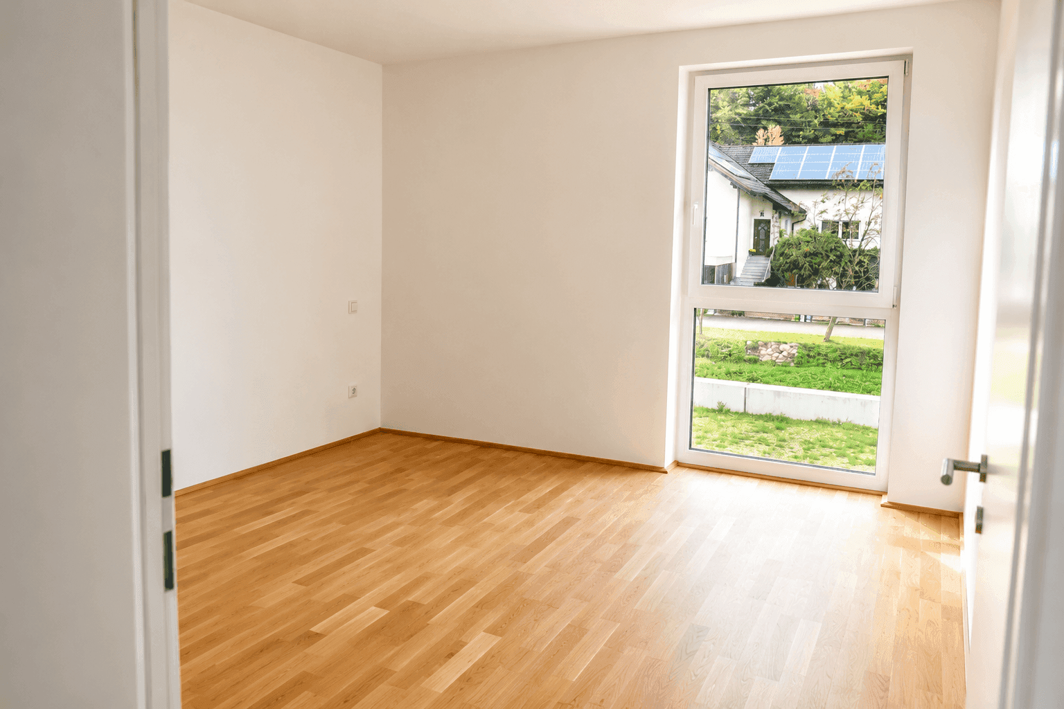 Leeres Schlafzimmer mit Parkettboden, großem Fenster und Blick ins Grüne.