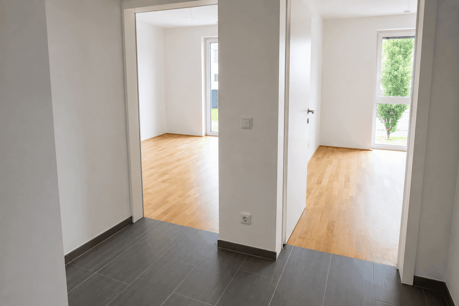 Heller Flur mit dunklem Fliesenboden, der zu angrenzenden Zimmern mit Parkett führt.