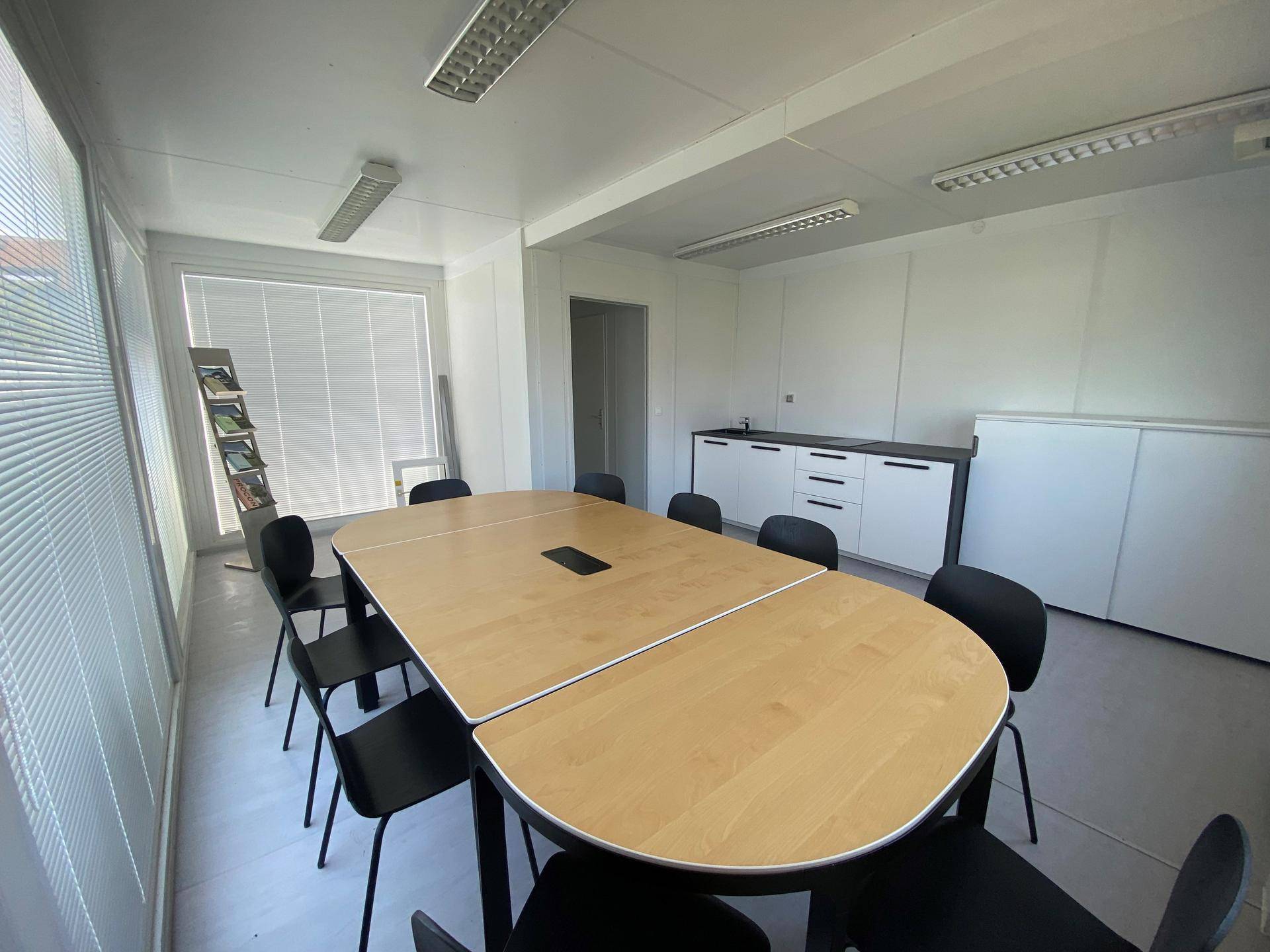 Funktionaler Büroraum mit ovalem Konferenztisch und Stühlen, ergänzt durch praktische Schränke und Jalousien an den Fenstern.