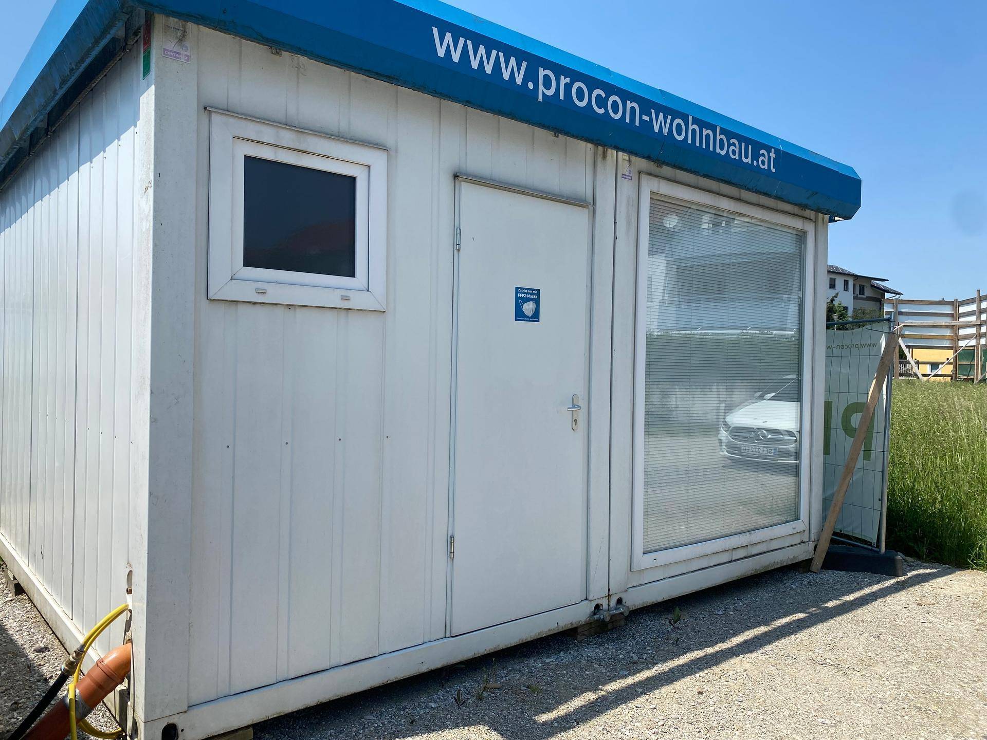 Weißes Containergebäude mit blauer Markise und großen Fenstern, dient als temporäres Büro oder Verkaufsraum.