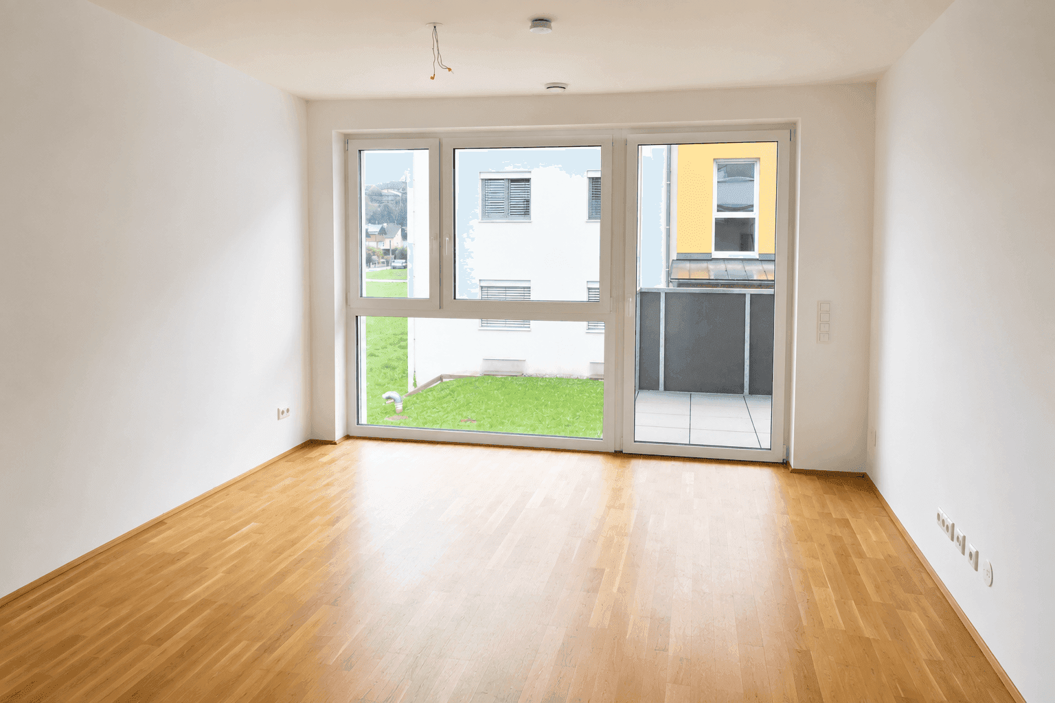 Heller, leerer Raum mit Parkettboden und großen Fenstern, die Zugang zu einer Terrasse bieten.