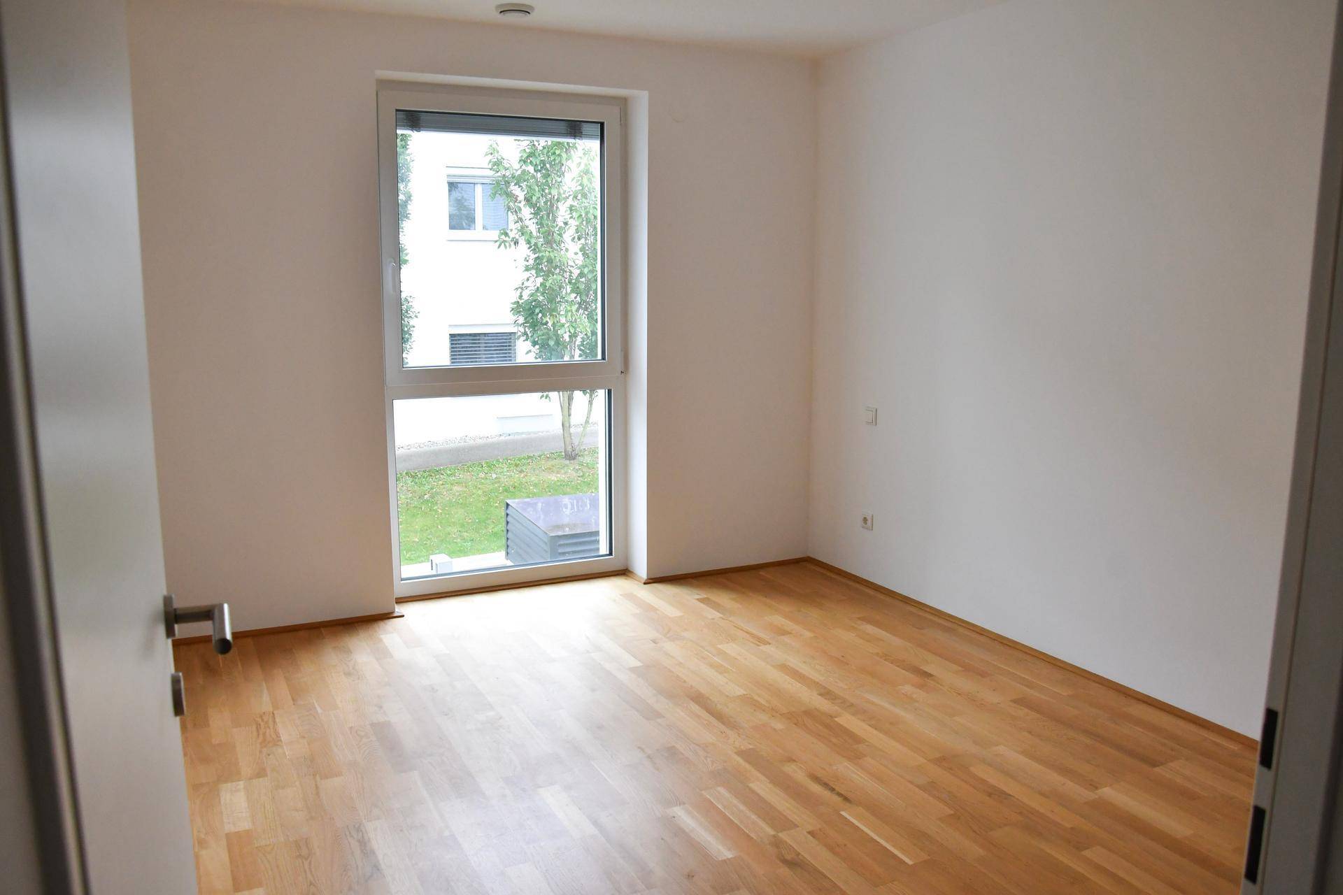 Helles, leeres Zimmer mit Holzboden und großem Fenster, das viel Tageslicht hereinlässt.
