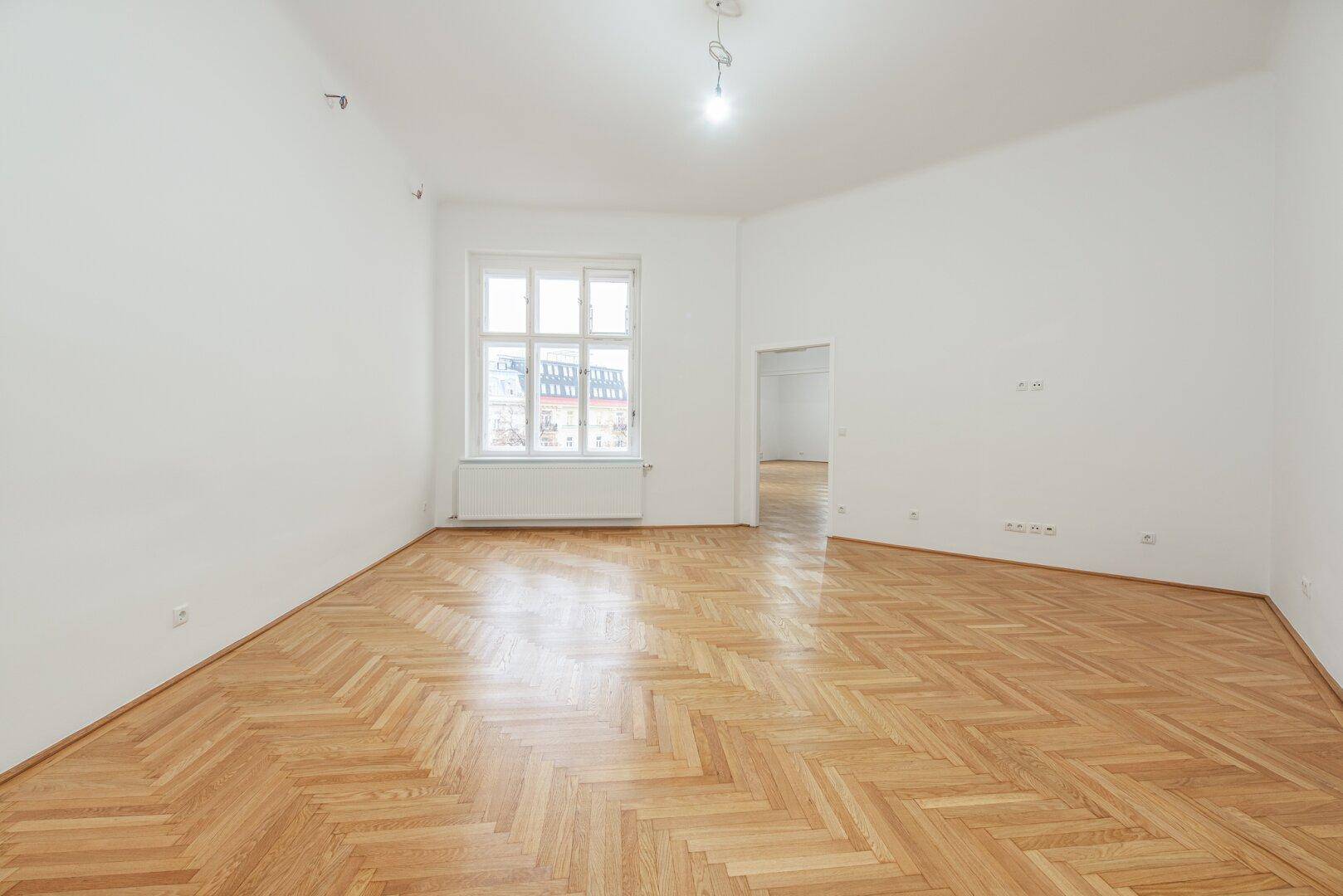 Heller Büroraum mit Fischgrätparkett, weißen Wänden und einem großen Fenster mit Stadtblick.