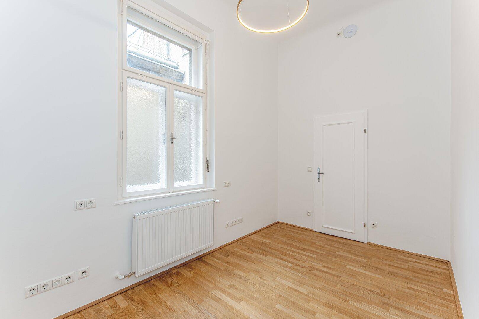 Geräumiger, heller Raum mit Parkettboden, hohen Decken und einem großen Fenster.