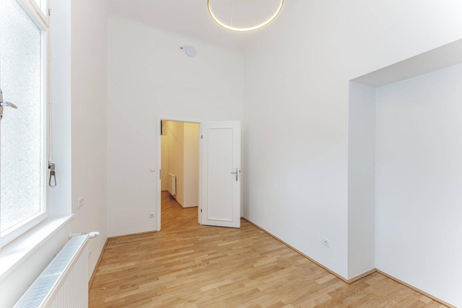 Leerer Raum mit hohen Decken, Parkettboden und Zugang zu einem weiteren Bereich.