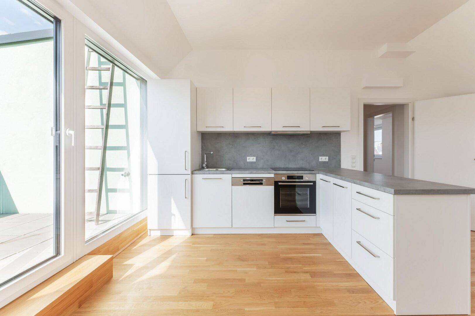 Moderne Küche mit weißen Schränken, Holzboden und Zugang zu einem Außenbereich durch eine große Glastür.