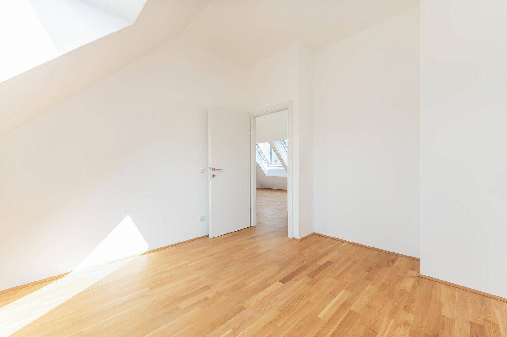 Heller Raum mit Holzboden und Dachschräge, der durch eine offene Tür einen Blick in einen weiteren Raum freigibt.