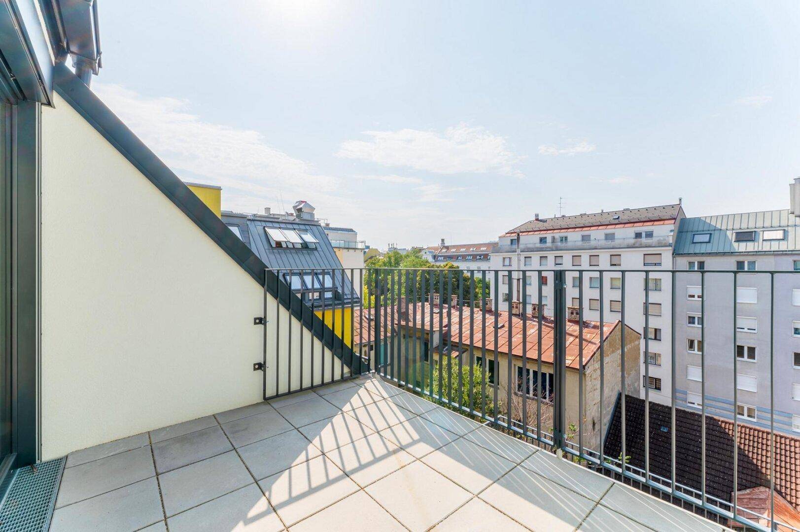 Sonniger Balkon mit Fliesenboden und Metallgeländer, der einen weiten Blick über die Dächer der Stadt bietet.
