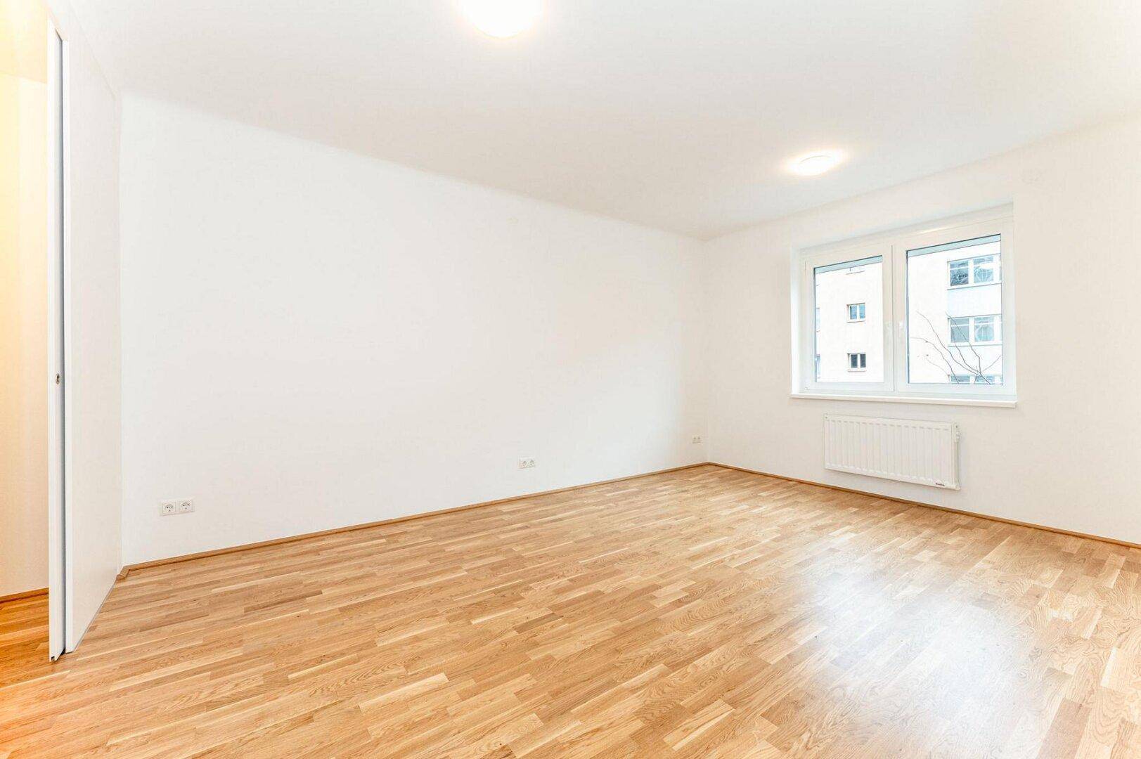 Helles Zimmer mit Parkettboden und großem Fenster, ideal als Wohn- oder Schlafzimmer nutzbar.