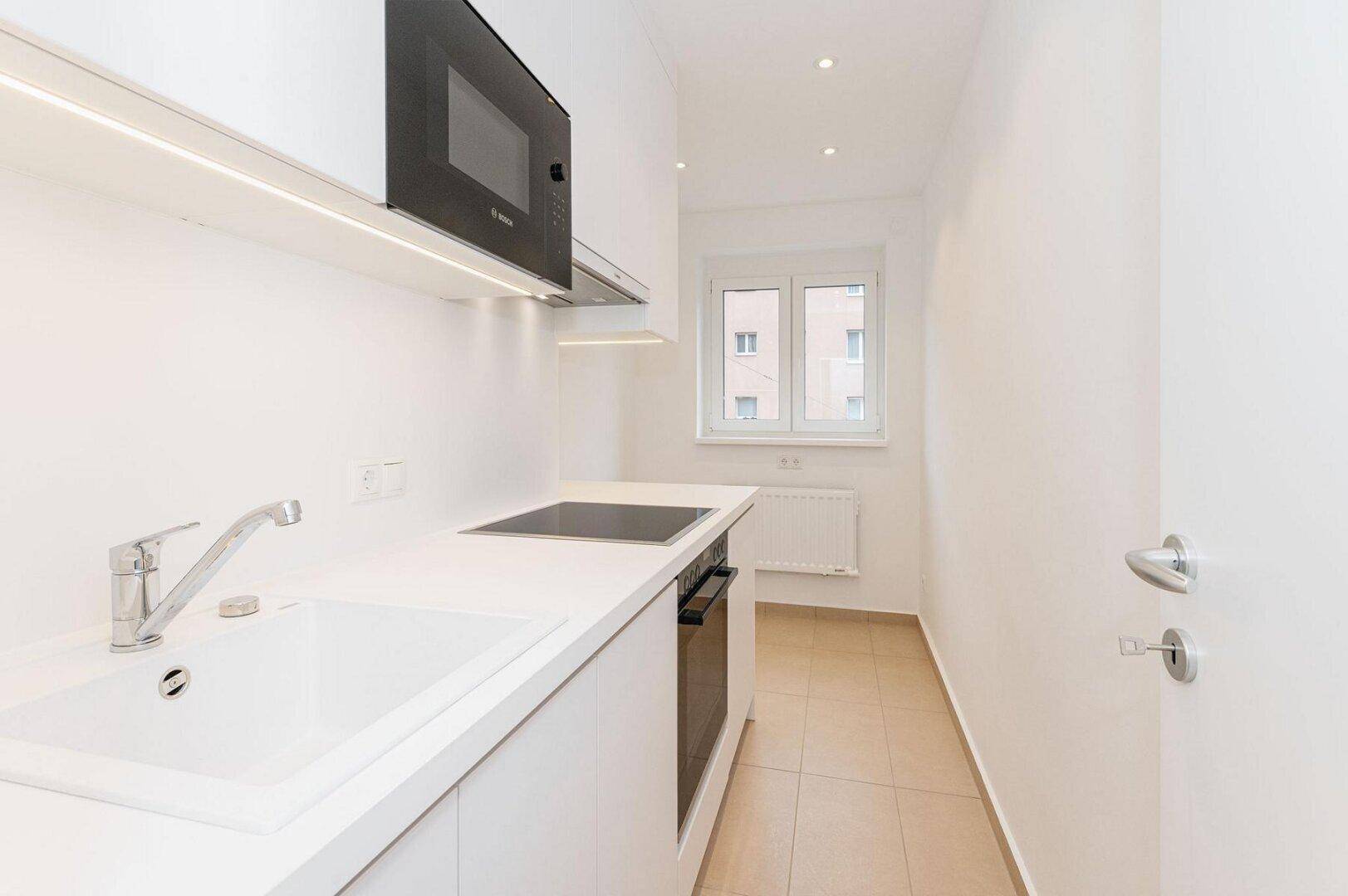 Moderne, schmale Küche mit weißen Schränken, Einbaugeräten und Fenster für Tageslicht.