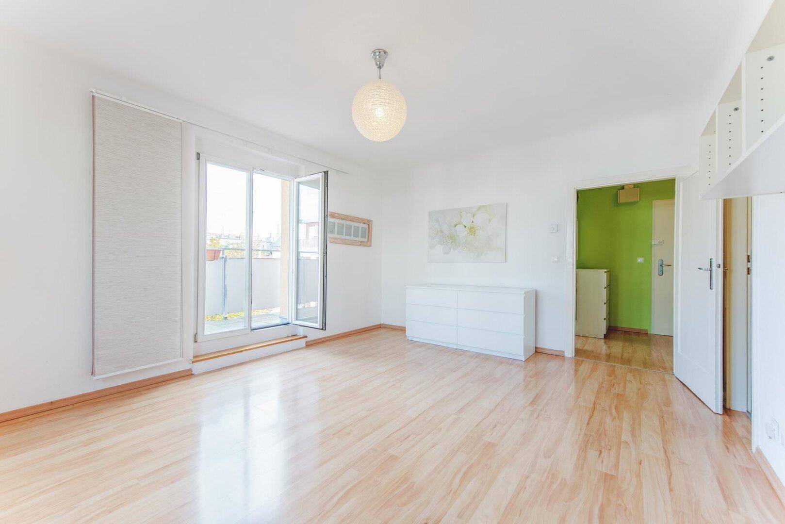 Geräumiges Wohnzimmer mit hellem Laminatboden, weißen Wänden und Zugang zu einem Balkon.