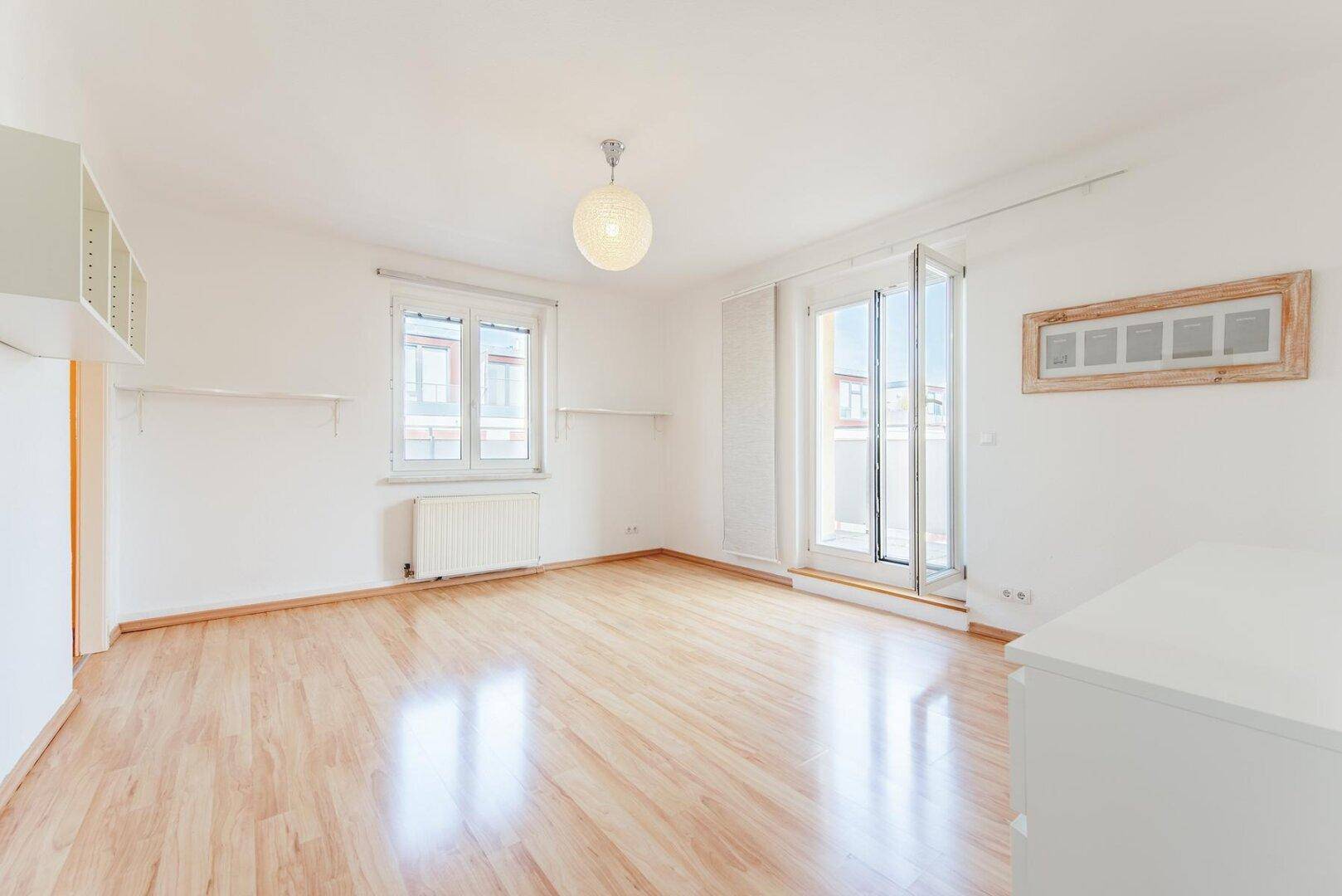 Helles Wohnzimmer mit Laminatboden, weißen Wänden und einem großen Fenster sowie Balkonzugang.