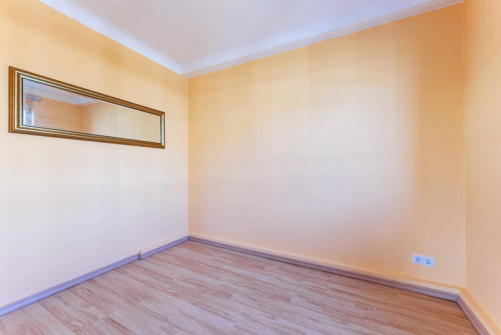 Kleines, helles Zimmer mit gelben Wänden, Laminatboden und einem dekorativen Wandspiegel.