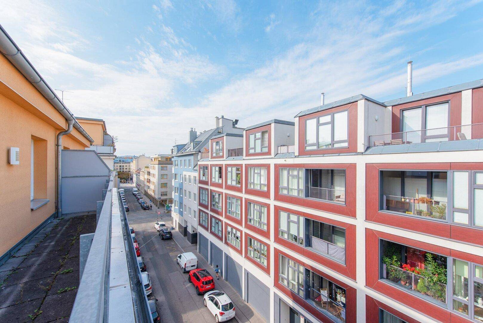Panoramablick auf die Straße mit modernen Gebäuden und parkenden Fahrzeugen unter blauem Himmel.