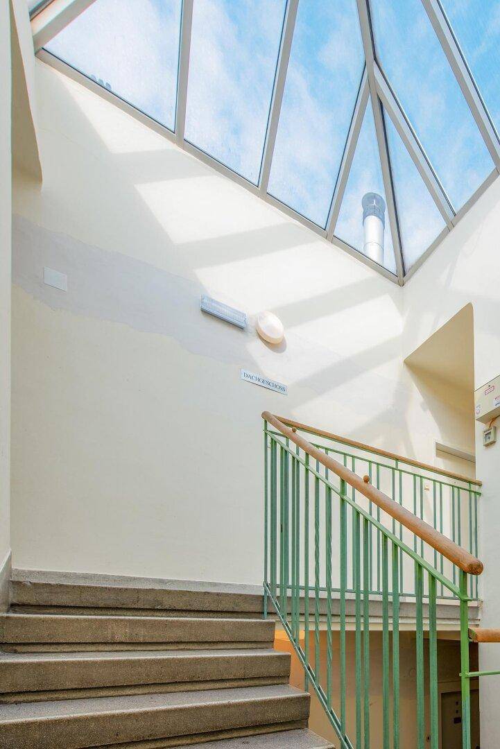 Helles Treppenhaus mit Steinstufen, grünem Geländer und einem großen Oberlicht für Tageslicht.