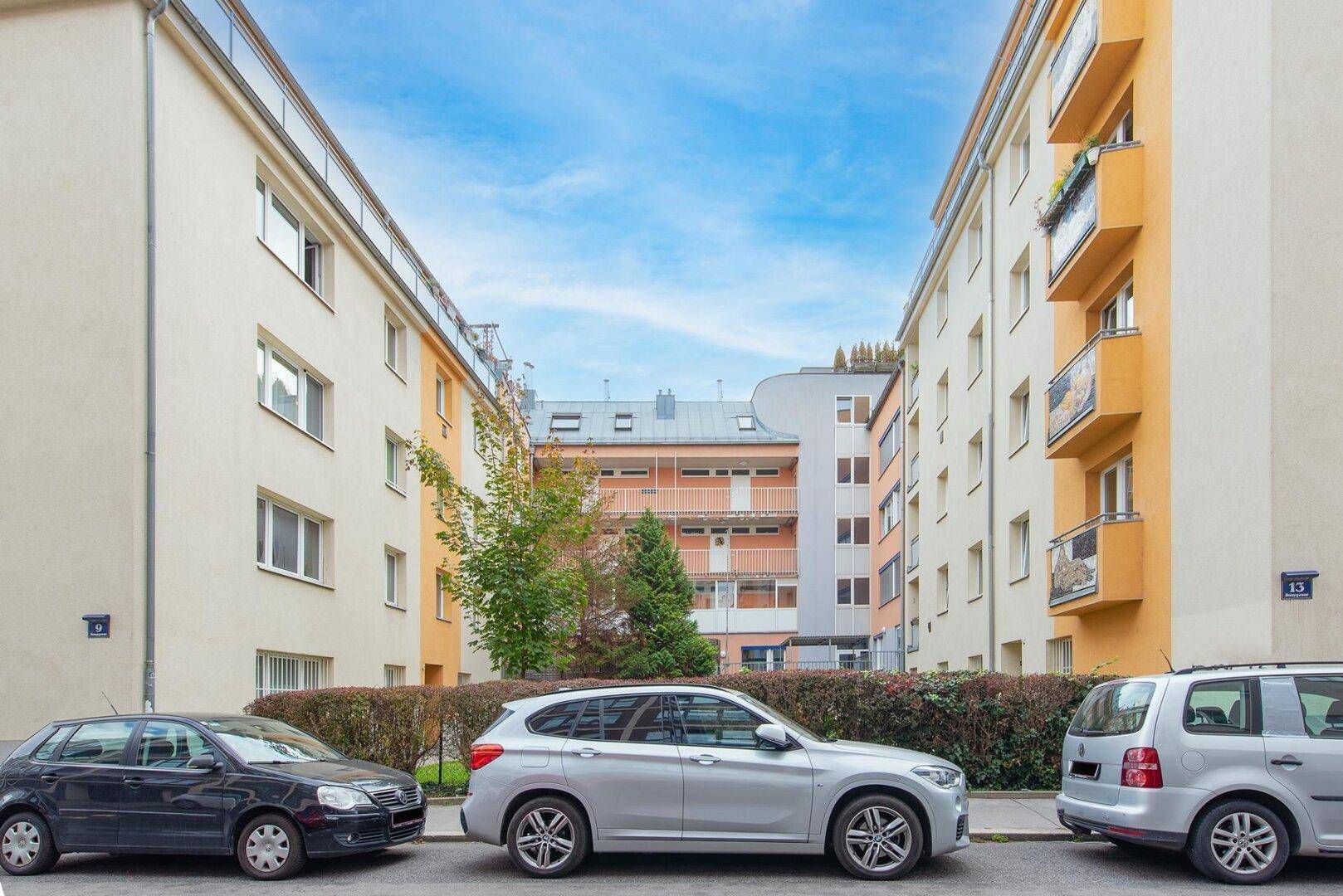 Blick auf die Straße mit parkenden Autos und Wohngebäuden im Hintergrund unter blauem Himmel.