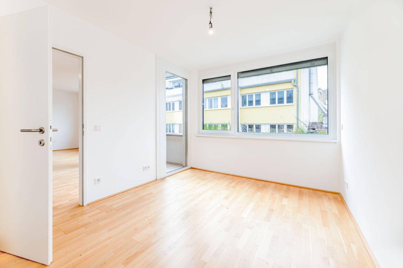 Helles Zimmer mit Parkettboden, großen Fenstern und Zugang zu einem kleinen Balkon.