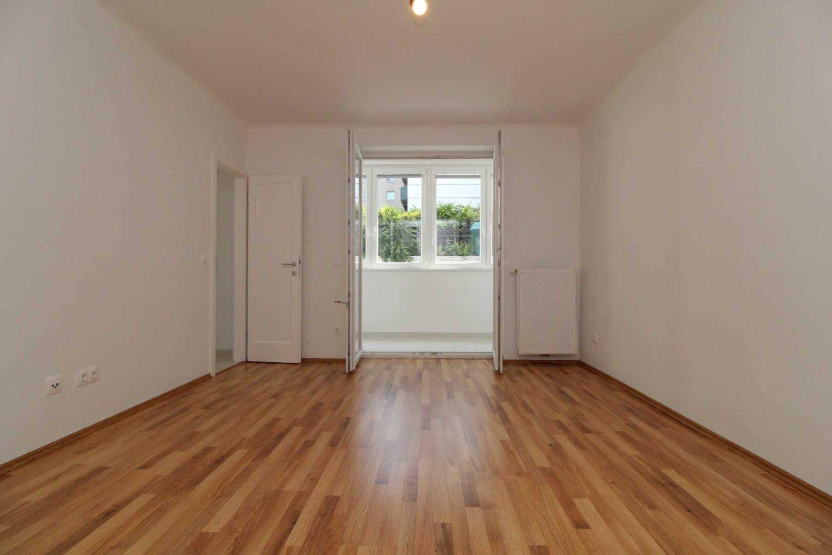 Geräumiges Zimmer mit hellem Parkettboden und geöffneter Balkontür, die viel Tageslicht hereinlässt.