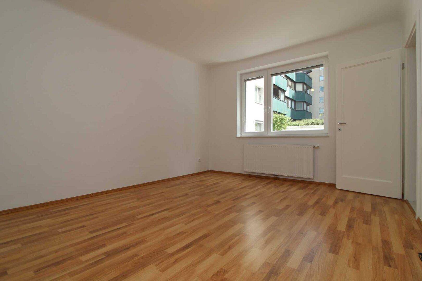 Ruhiges Schlafzimmer mit Parkettboden und Fenster, das einen Blick auf die Nachbarschaft bietet.