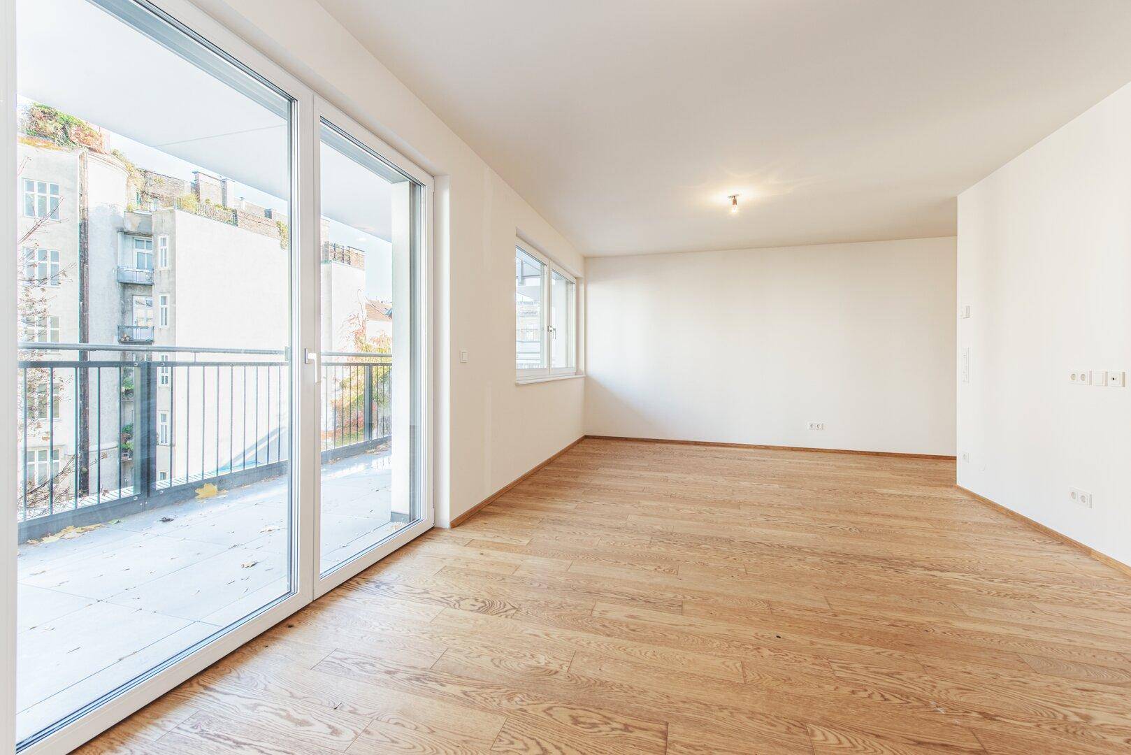 Geräumiger Wohnbereich mit hellem Parkettboden und großen Schiebetüren zum Balkon.