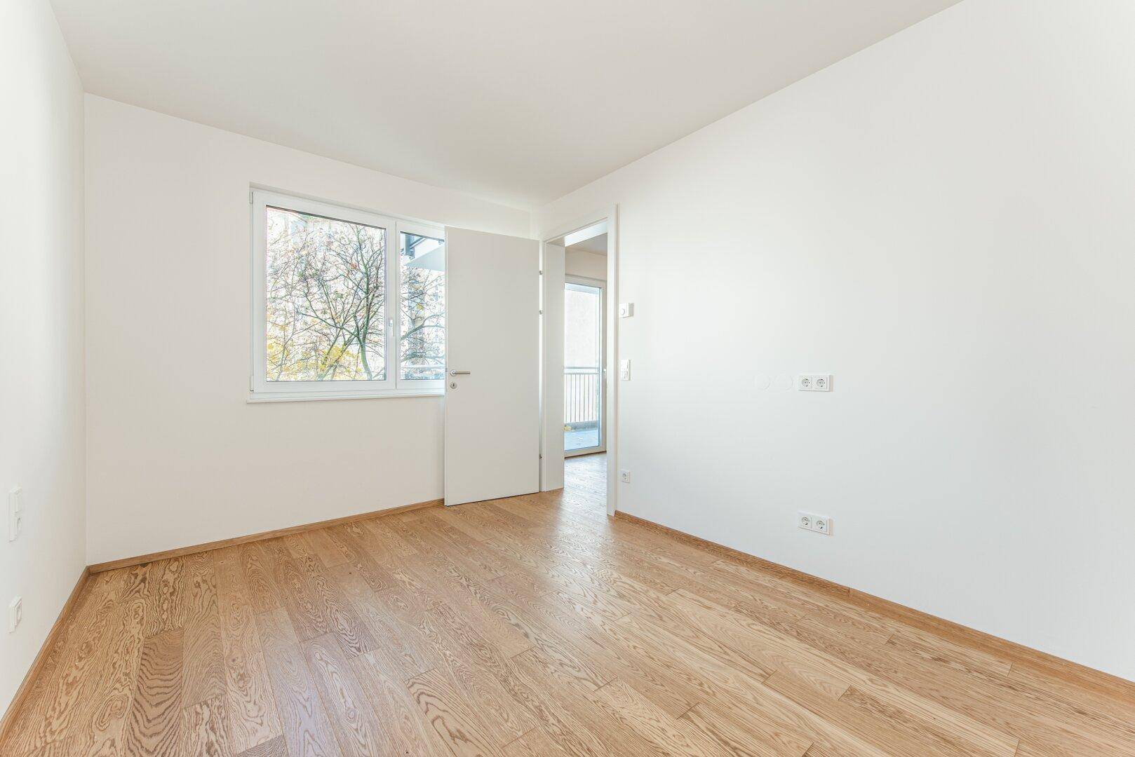 Helles Schlafzimmer mit Parkettboden und Fenster mit Blick ins Grüne.