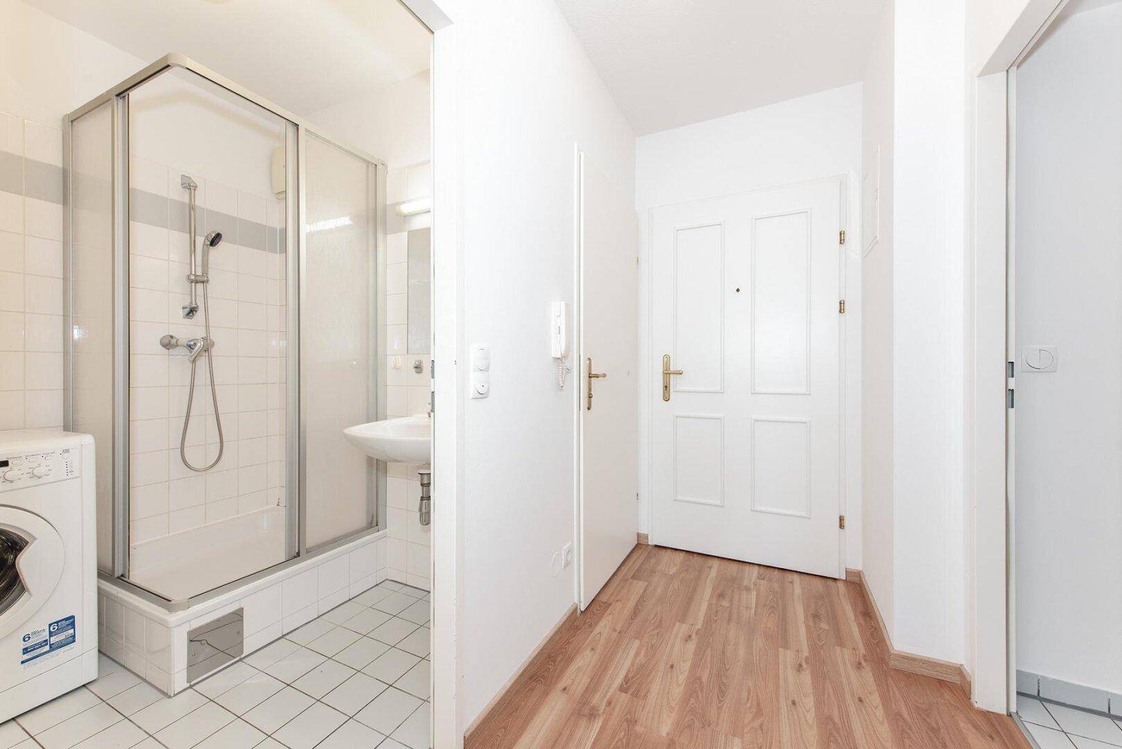 Kompaktes Badezimmer mit Duschkabine, Waschbecken und integrierter Waschmaschine im Eingangsbereich.