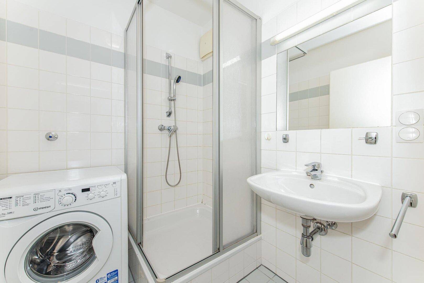 Helles Badezimmer mit Duschkabine, Waschbecken und Waschmaschine, alles in weiß gefliest.
