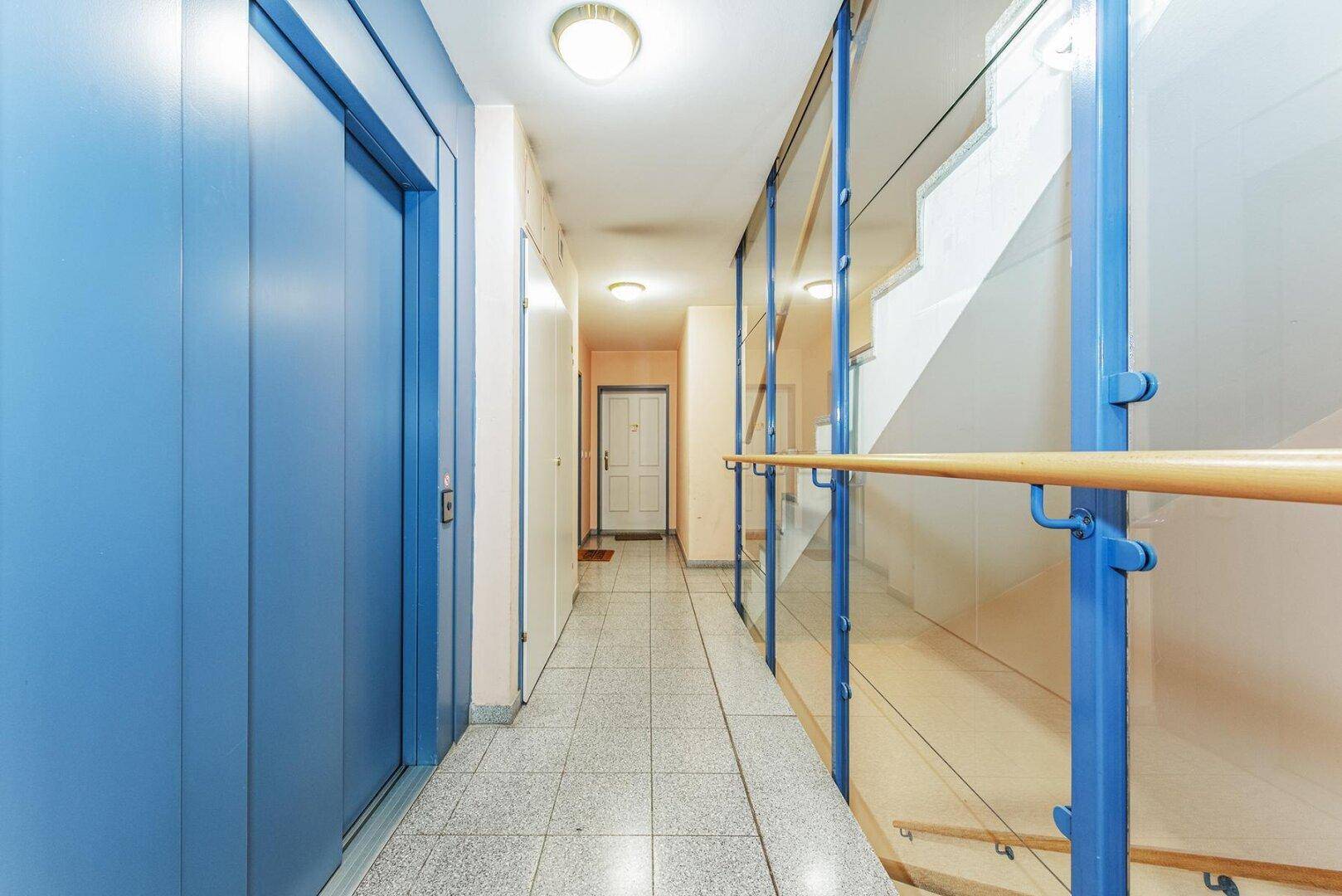Heller Hausflur mit Aufzug und Treppenhaus, ausgestattet mit blauen Akzenten und Fliesenboden.