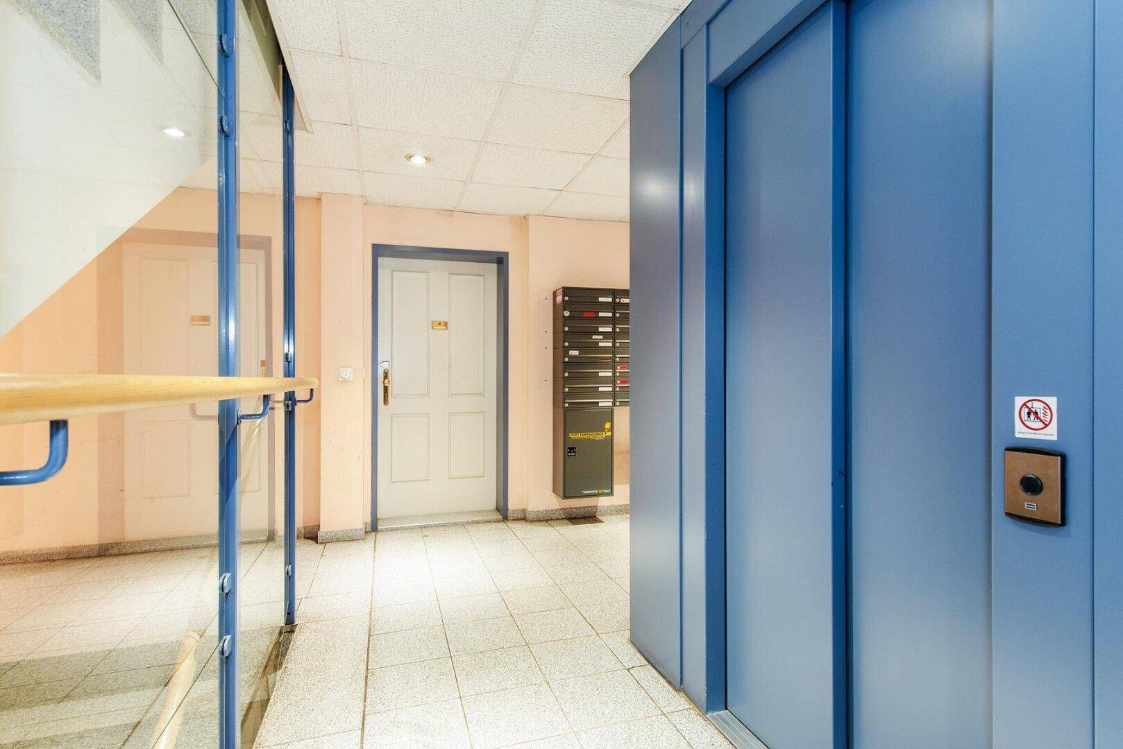 Moderner Hausflur mit blauem Aufzug und Briefkästen, führt zum Treppenhaus.