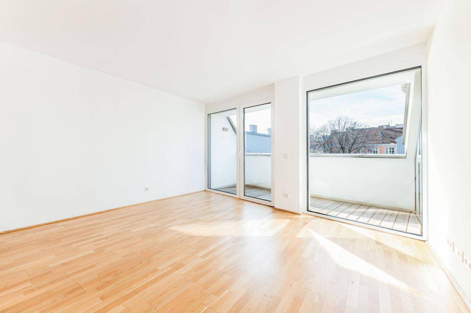 Helles, leeres Wohnzimmer mit Parkettboden und großen Fenstern, die auf einen Balkon führen.
