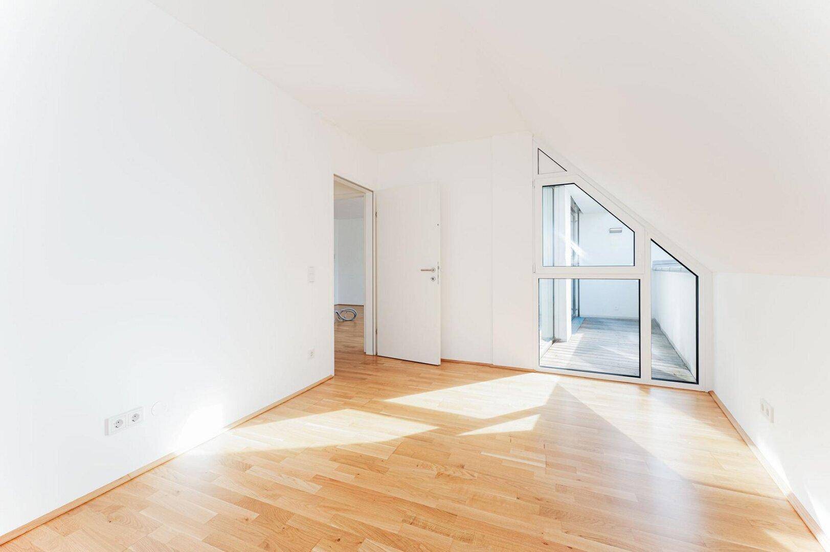 Helles Schlafzimmer mit Parkettboden, Dachschräge und Zugang zu einem sonnigen Balkon.