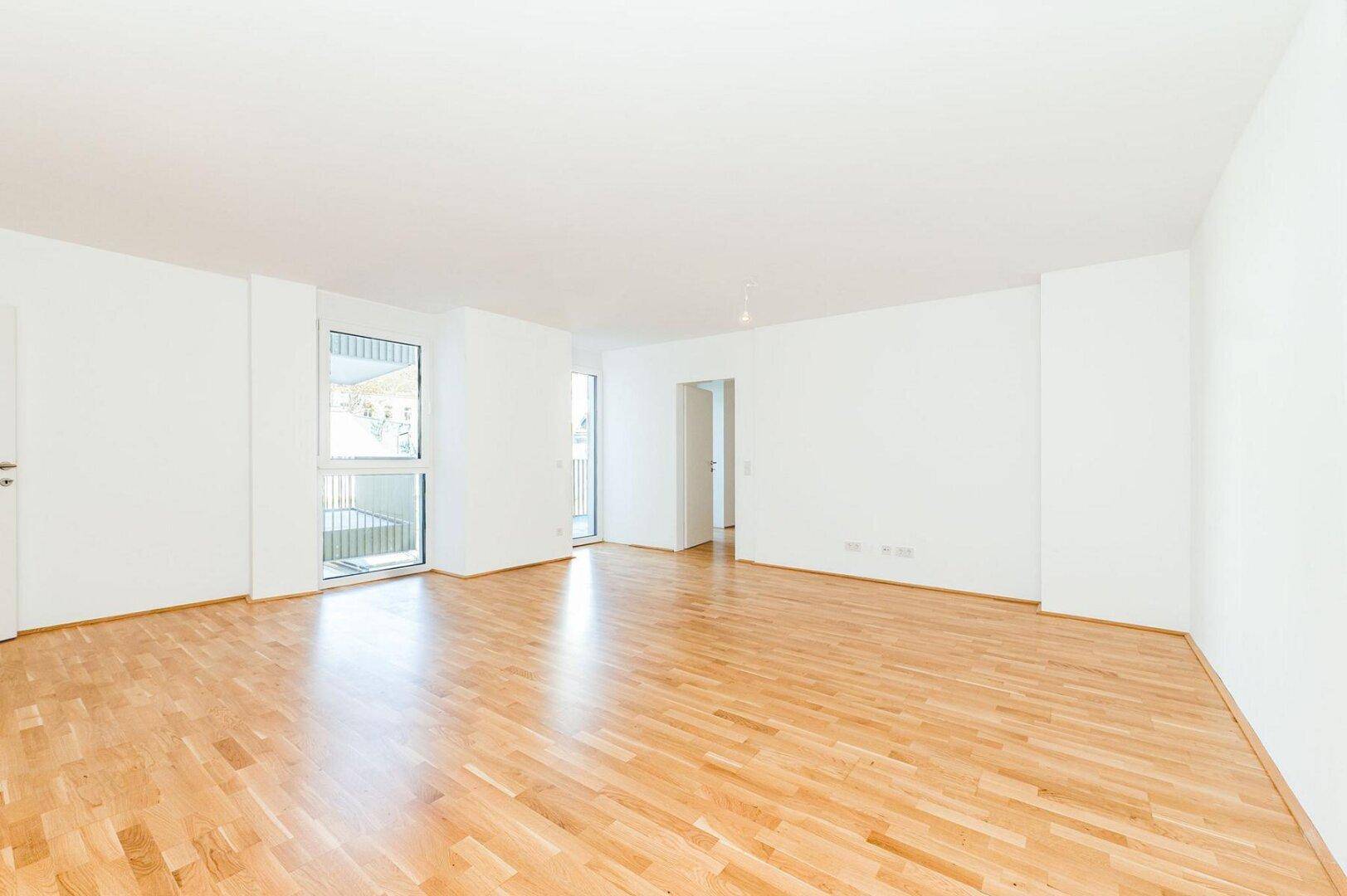 Geräumiges, leeres Wohnzimmer mit hellem Parkettboden und großen Fenstern für viel Tageslicht.