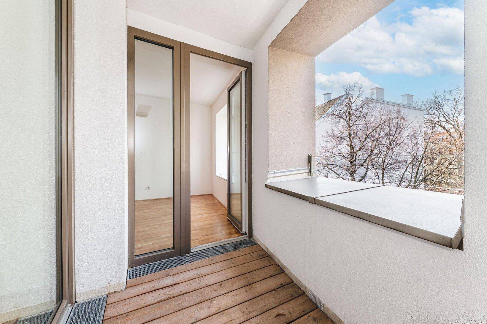 Heller Balkon mit Holzboden und Blick auf die umliegenden Gebäude und Bäume.