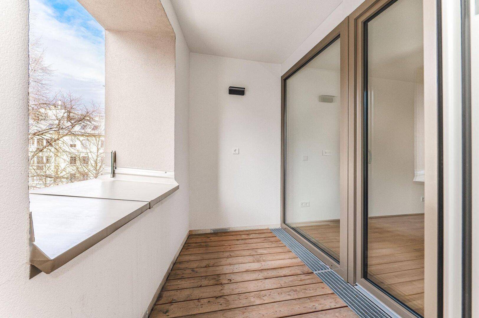 Geräumiger Balkon mit Holzdielen und Fensterfront, die viel Tageslicht hereinlässt.