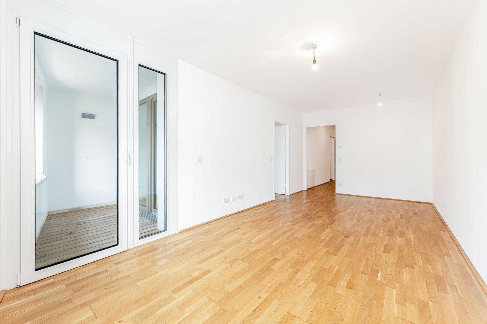 Helles, leeres Wohnzimmer mit Parkettboden und großen Fenstern zum Balkon.