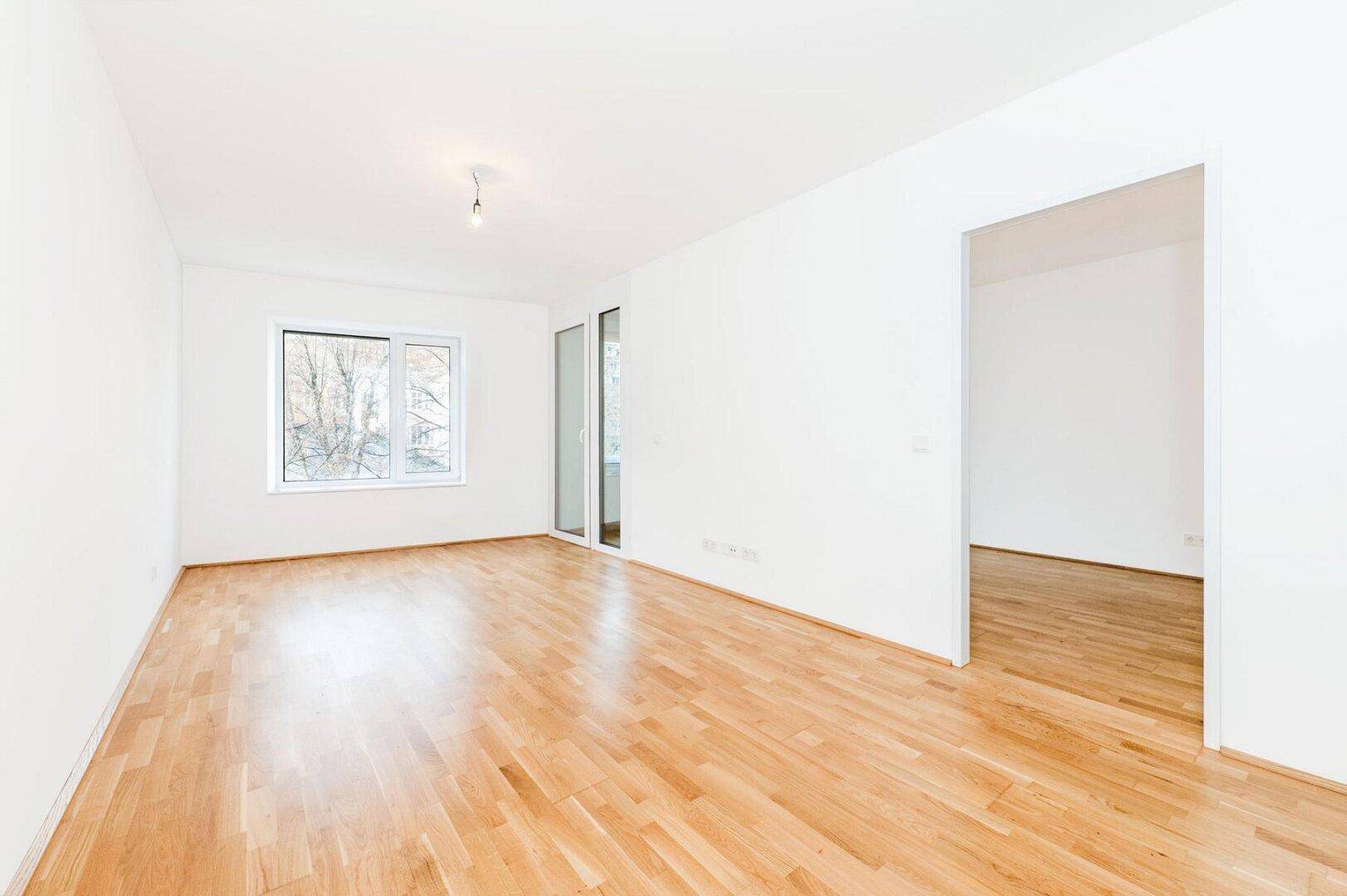Geräumiges Wohnzimmer mit hellem Parkettboden und einem großen Fenster, das viel Licht spendet.