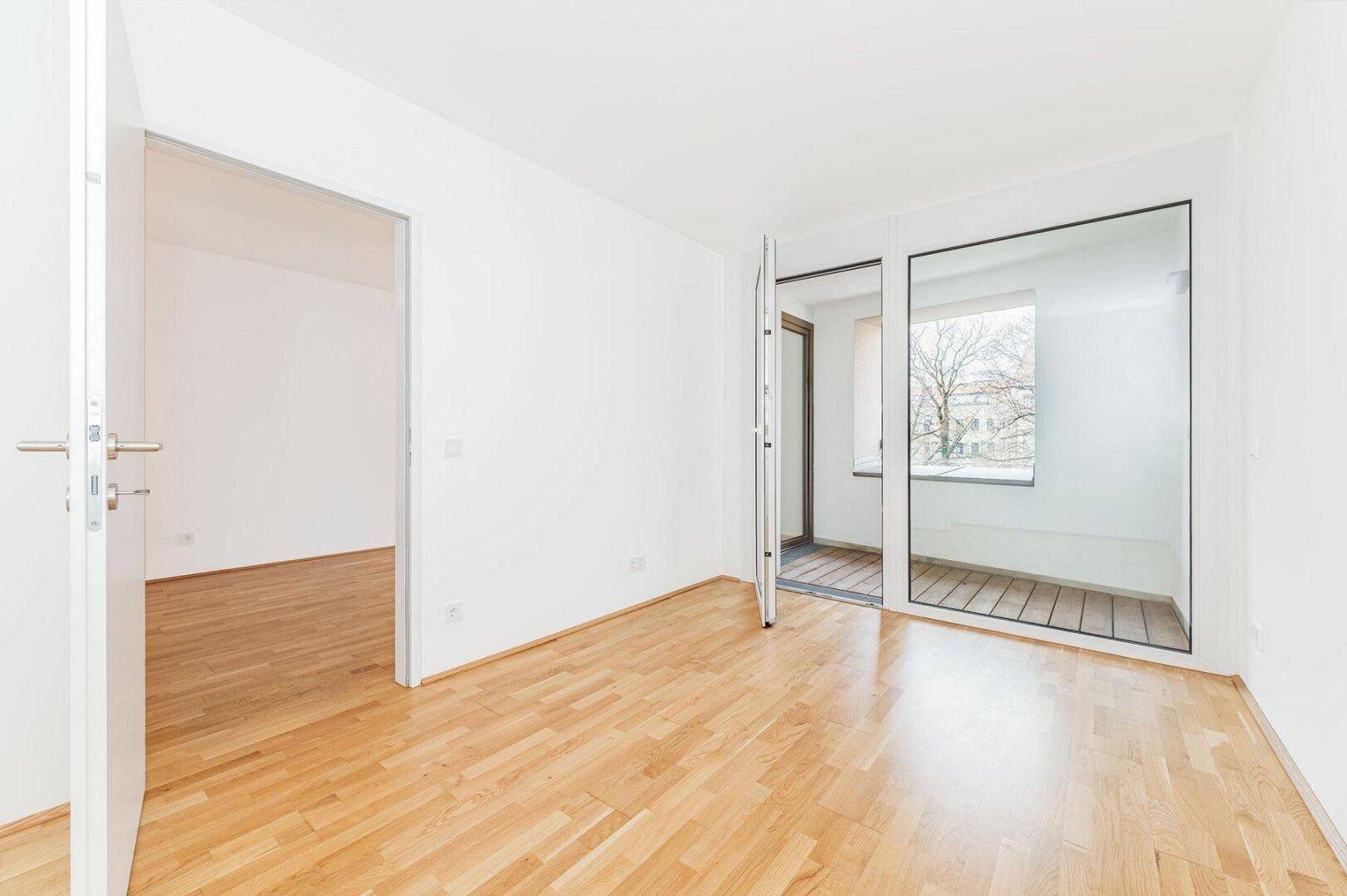 Schlafzimmer mit hellem Parkettboden und Zugang zu einem Balkon durch große Glastüren.