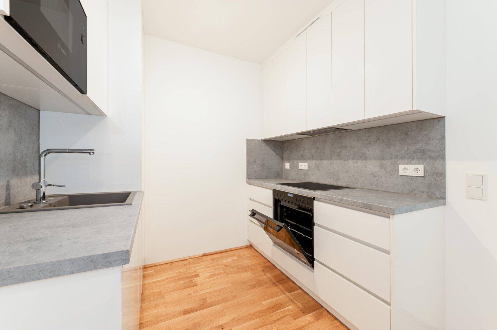 Helle Einbauküche mit geöffnetem Backofen, weißen Schränken und grauer Arbeitsplatte.