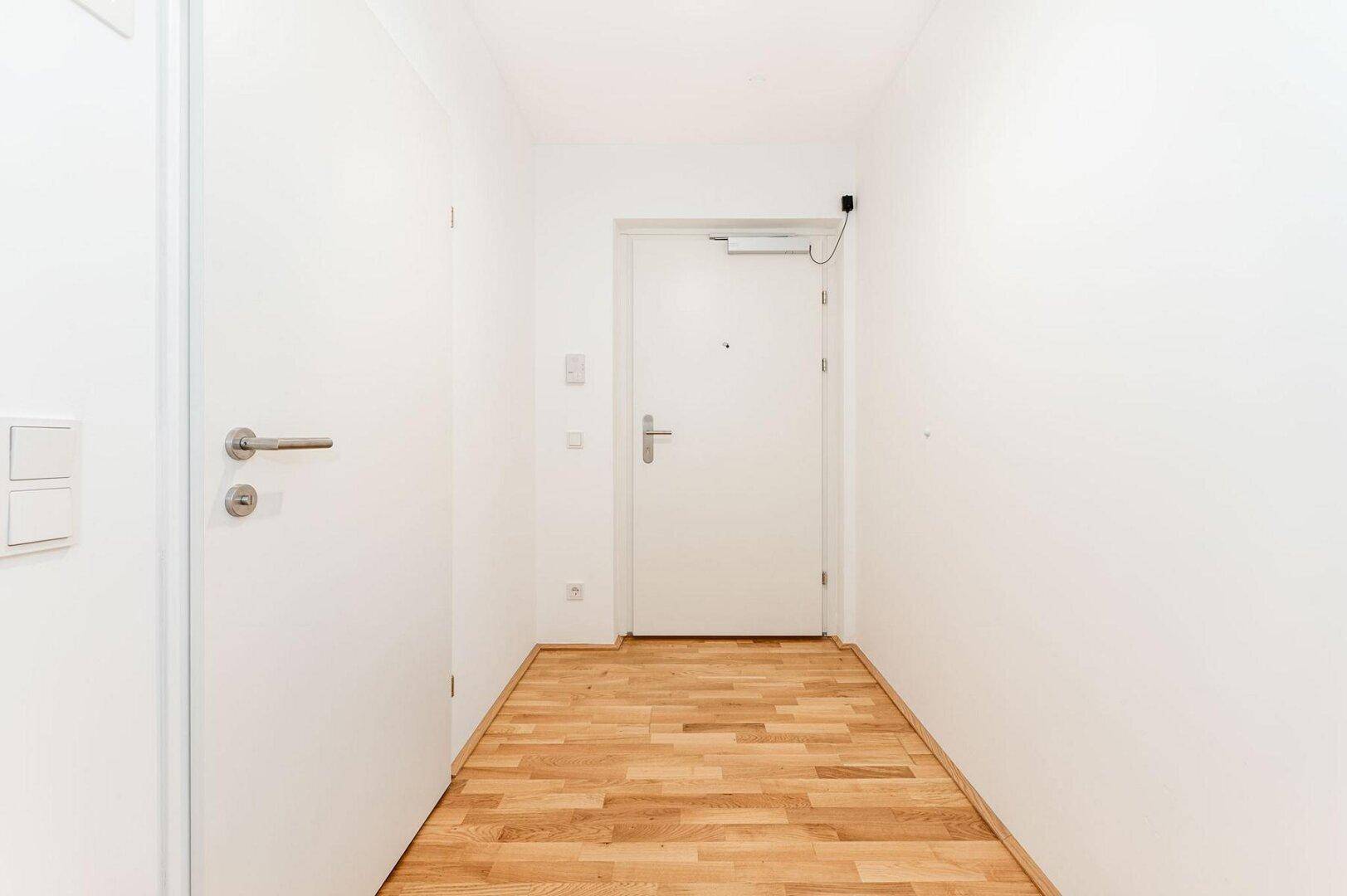 Heller Eingangsbereich mit Parkettboden und weißer Eingangstür, die in die Wohnung führt.