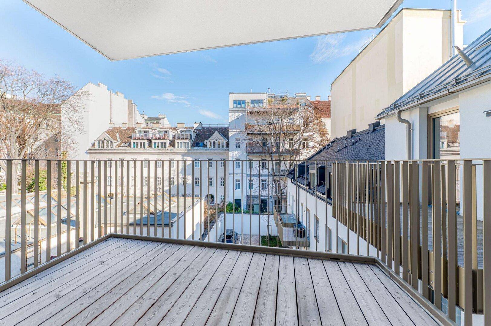 Geräumiger Balkon mit Holzdielen und Blick auf die Dächer der umliegenden Gebäude unter blauem Himmel.