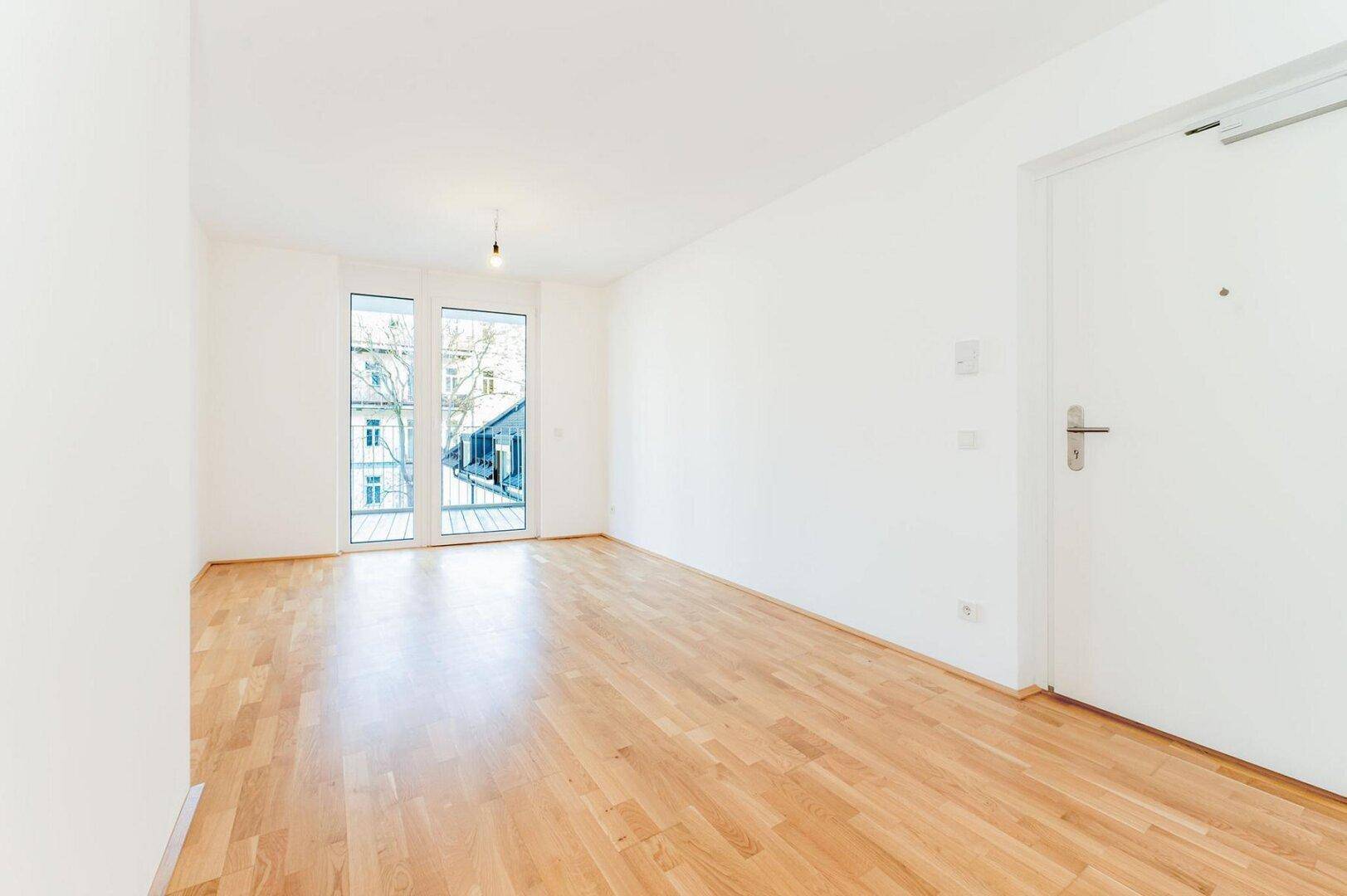 Heller, leerer Raum mit hellem Parkettboden und großen Fenstertüren, die auf einen Balkon führen.