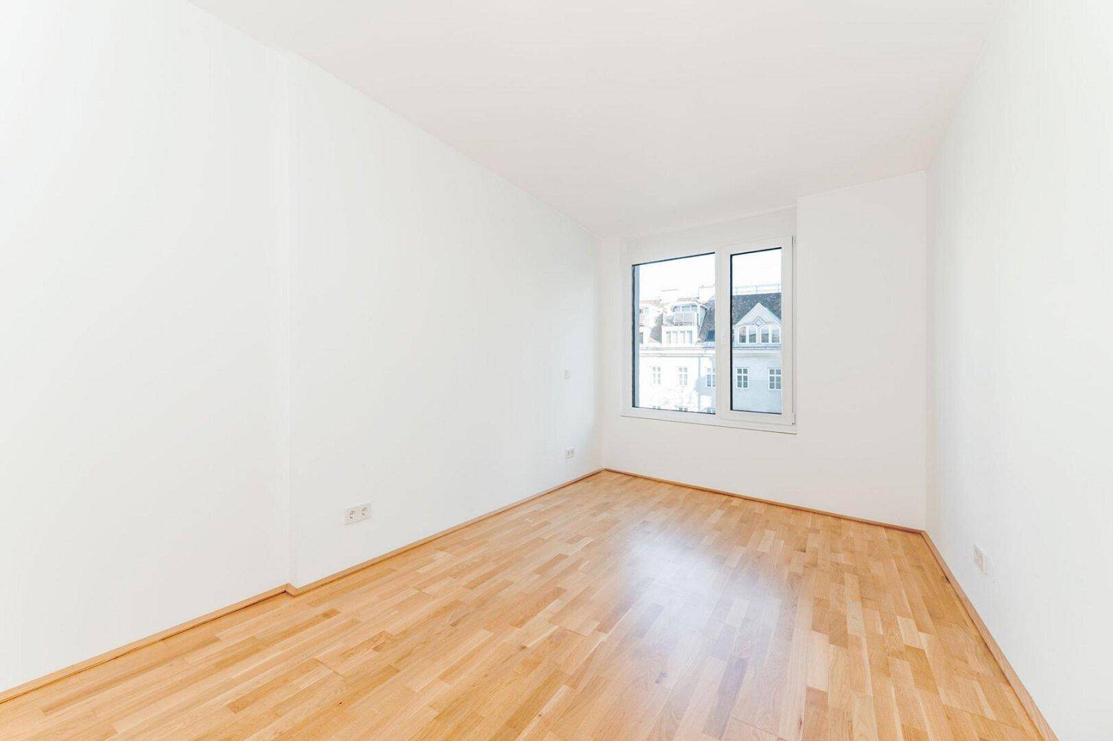Helles Schlafzimmer mit Parkettboden und großem Fenster, das viel Tageslicht hereinlässt.