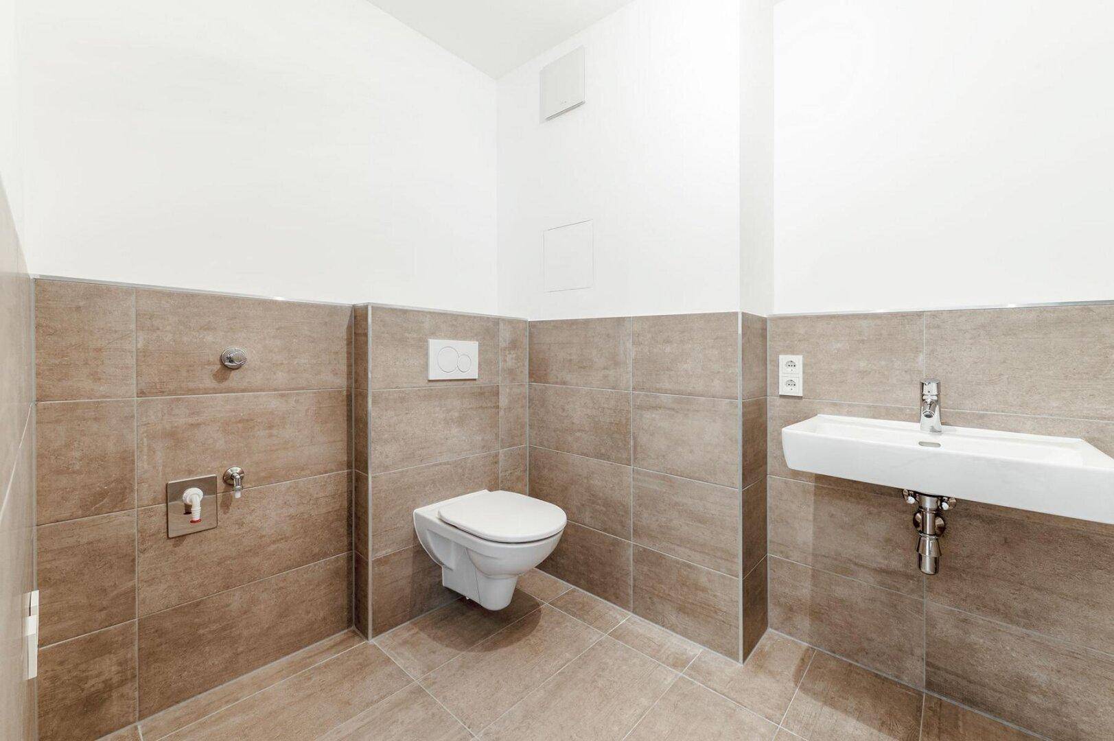 Separates WC mit braunen Fliesen, wandhängender Toilette und einem kleinen Handwaschbecken.