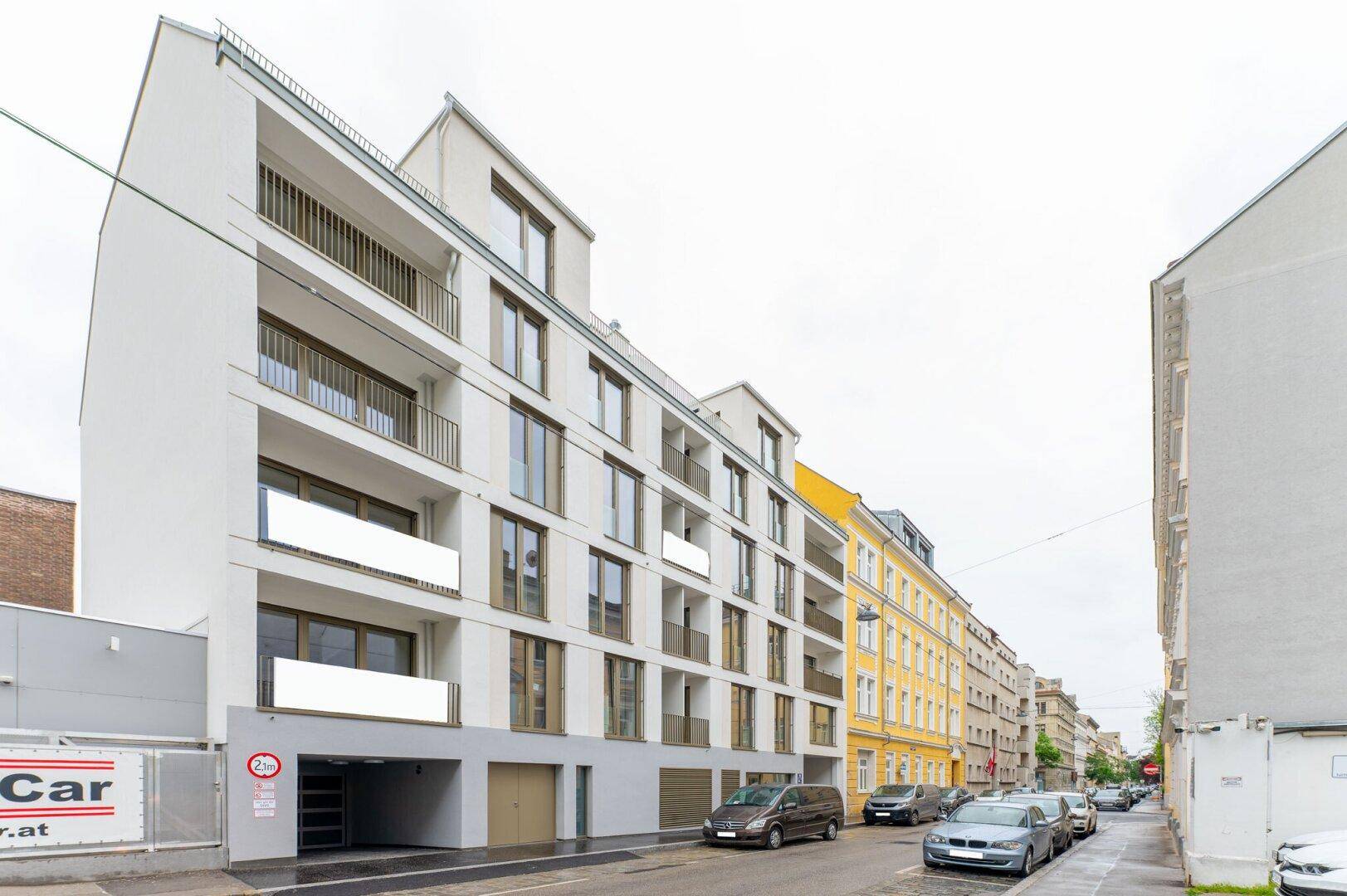 Ansicht des modernen Neubaus von der Straße mit parkenden Autos und angrenzenden Gebäuden.
