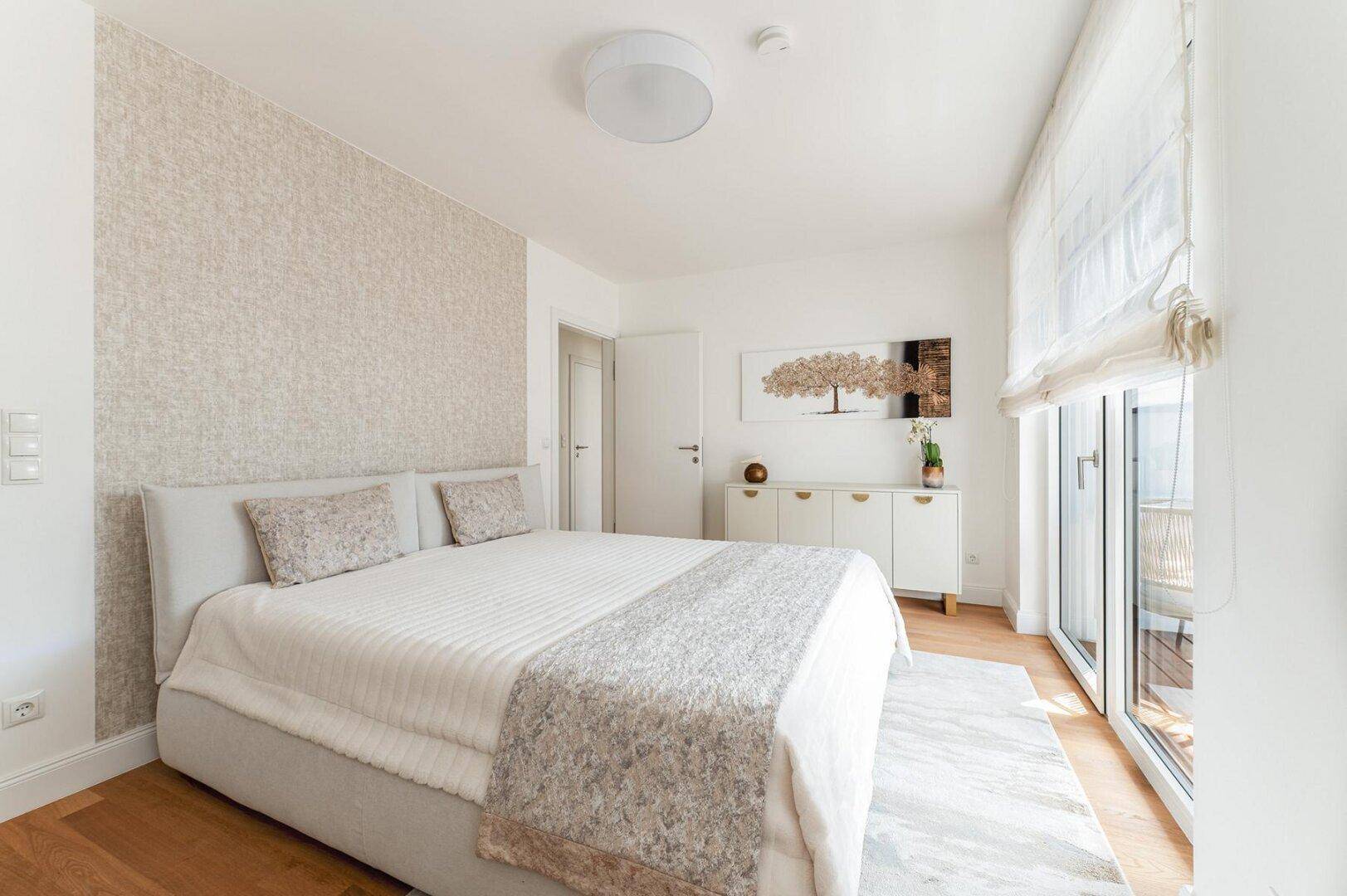 Helles Schlafzimmer mit Doppelbett, Holzboden und Zugang zum Balkon durch große Fenster.