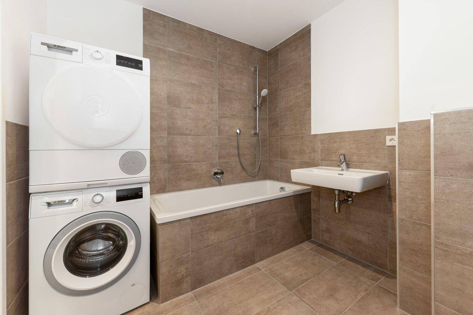 Modernes Badezimmer mit Badewanne, Waschmaschine und Trockner, ausgestattet mit beigefarbenen Fliesen.