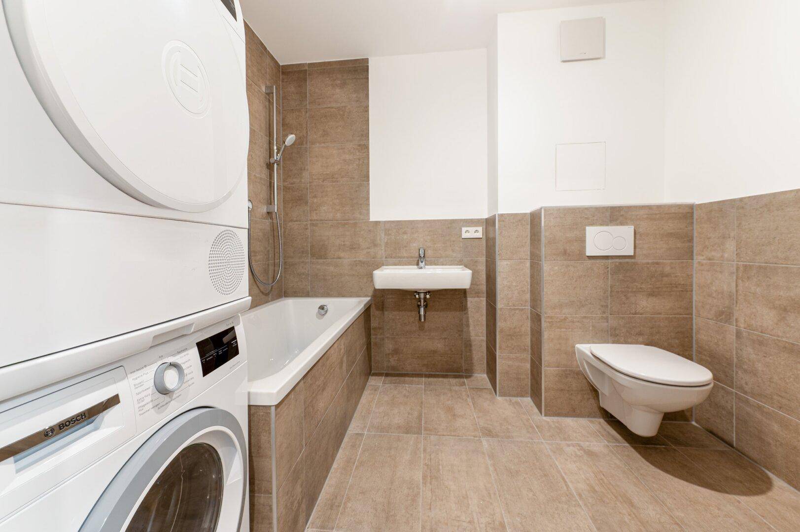 Helles Badezimmer mit Badewanne, Waschmaschine, Trockner und wandhängendem WC in neutralen Farbtönen.