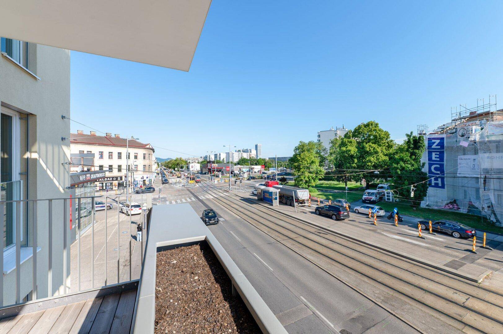 Weitläufiger Blick von einem Balkon auf eine städtische Kreuzung mit Straßenbahnschienen und Gebäuden.